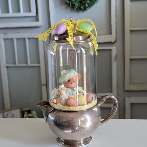 May include: A silver creamer with a glass cloche on top. Inside the cloche is a teddy bear wearing a blue beret and a white dress. The teddy bear is surrounded by pastel-colored sprinkles and candy.