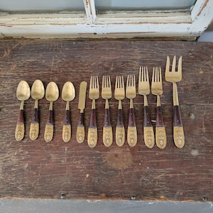 May include: An assortment of antique cutlery, including spoons, forks, and a butter knife, displayed on a wooden surface. The utensils feature brass heads and dark brown wooden handles with decorative brass detailing. The set includes various sizes of forks and spoons.