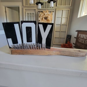 May include: A wooden brush with black and white letters spelling out the word "JOY" on top of it. The letters are propped up on the brush bristles.