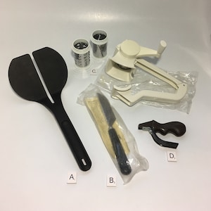 May include: A set of kitchen tools including a black spatula, a white grater with a handle, a black knife in a plastic bag, and a brown and black hand-held tool labeled "D".