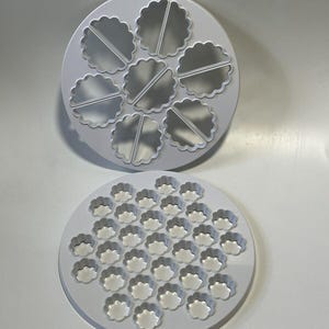 Puede incluir: Dos moldes circulares blancos de plástico con bordes festoneados. El molde superior presenta siete cavidades grandes en forma de flor, cada una dividida por una barra recta. El molde inferior contiene treinta y siete pequeñas hendiduras florales.