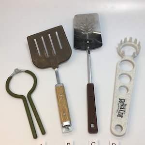 May include: A collection of kitchen utensils, including a slotted spatula, a solid spatula, a pasta measuring tool, and a bottle opener. The utensils have wooden, brown, and stainless steel handles. The pasta tool has the text "Franco Rinaldi Pasta Sauce".