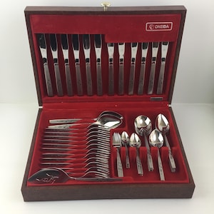 May include: A vintage Oneida silverware set in a wooden case with a red velvet interior. The set includes knives, forks, spoons, and serving utensils. The silverware has a floral design on the handles. The case is brown with a gold clasp.