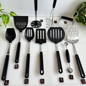 May include: A collection of kitchen utensils including a black spatula, a slotted turner, a potato masher, a wire whisk, a pastry blender, a melon baller, and a metal spatula. Each tool features a black handle and is labeled with a small letter tile.
