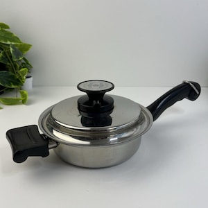 Op de afbeelding: Een zilveren pan met een zwarte handgreep en deksel. Het deksel heeft een zwarte knop.