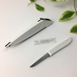 Op de afbeelding: Een witte en zilveren schilmes met een bijpassende schede en een kleine productinformatiekaart. Het mes heeft een zilveren lemmet en een witte handgreep. De schede is wit en hoekig. De productkaart bevat tekst.