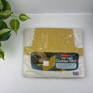 以下が含まれることがあります： 透明なプラスチック包装に入った黄色のラバーメイド製ツインシンクディバイダーマット。ラベルにはRubbermaid TWIN SINK Divider Matの文字があり、食器やシンクを保護すると記載されています。サイズは27.9cm x 33cmです。