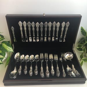 May include: A silver-toned flatware set arranged in a dark wooden storage case with a black velvet interior. The collection includes various spoons, forks, and knives, each featuring an ornate, embossed handle design with intricate floral detailing.