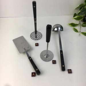 May include: A collection of vintage kitchen utensils, including a spatula, potato masher, meat tenderizer, and ladle. Each tool has a black handle and a metal working end. The tools are arranged on a white surface.