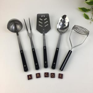 May include: A set of five stainless steel kitchen utensils with black handles. Includes a ladle, fork, spatula, slotted spoon, and potato masher. Scrabble tiles spell out A, B, C, D, and E.