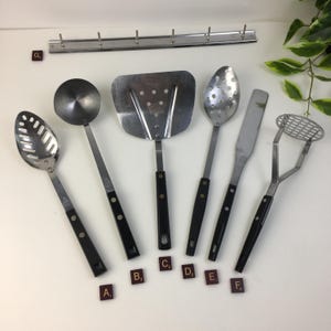 Puede incluir: Un juego de utensilios de cocina vintage, que incluye una cuchara ranurada, un cucharón, una espátula y un machacador de patatas. Los utensilios tienen cabezas de acero inoxidable y mangos negros. Encima de los utensilios hay un estante metálico con ganchos.
