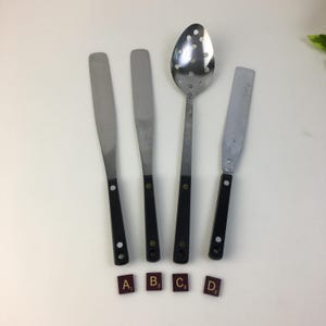 Puede incluir: Cuatro utensilios de cocina: dos espátulas, una cuchara ranurada y una espátula con mango negro. Los utensilios son de acero inoxidable. Cuatro fichas de Scrabble deletrean A, B, C y D.
