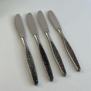 May include: A set of four silver-toned stainless steel dinner knives arranged in a row. Each knife features a smooth, rounded blade and a textured handle decorated with an intricate, embossed floral pattern covering the entire length of the handle.