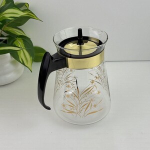 Puede incluir: Cafetera de cristal transparente con asa y tapa negras. La cafetera presenta una banda dorada y un diseño floral dorado. La cafetera es de estilo vintage.