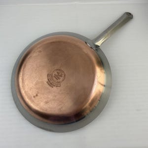 Könnte beinhalten: Eine Revere Ware Bratpfanne mit Kupferboden und Edelstahl-Außenseite und Griff. Die Pfanne hat eine runde Form mit einem Durchmesser von ca. 25 cm. Der Griff ist silberfarben und nach oben abgewinkelt.