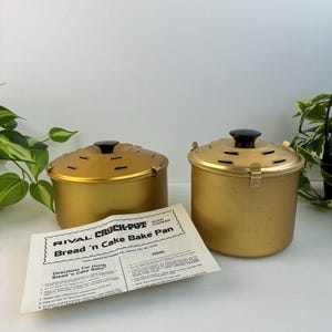 May include: Two gold-coloured metal Rival Crock-Pot Bread 'n Cake Bake pans with black knobs on the lids. Each lid features ventilation slots and metal clips. An instruction sheet titled Rival Crock-Pot Bread 'n Cake Bake Pan is placed in front.