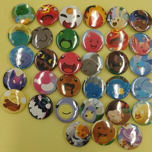 May include: A collection of round, colorful pinback buttons with various cartoon character designs. The buttons feature a variety of colors, including green, red, blue, and yellow, with detailed illustrations of creatures and faces. The buttons are arranged on a yellow surface.