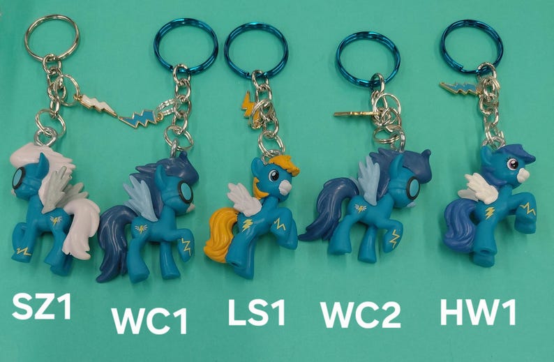 K&ouml;nnte beinhalten: F&uuml;nf blaue und t&uuml;rkisfarbene Pony-Schl&uuml;sselanh&auml;nger mit wei&szlig;en Fl&uuml;geln und gelben Akzenten. Jeder Schl&uuml;sselanh&auml;nger hat eine silberne Kette und einen blauen Ring. Die Schl&uuml;sselanh&auml;nger sind mit SZ1, WC1, LS1, WC2 und HW1 beschriftet.