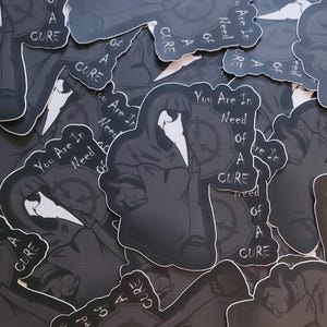 May include: A collection of black and white stickers with the text "You Are In Need Of A Cure" and a graphic of a figure in a hooded robe. The stickers are printed on a white background.