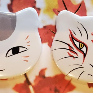 Op de afbeelding: Twee witte, katvormige knopen van keramiek. De ene knop heeft een witte kat met oranje en grijze markeringen. De andere knop heeft een witte kat met rode markeringen rond de ogen en een rode draaikolk op het voorhoofd.