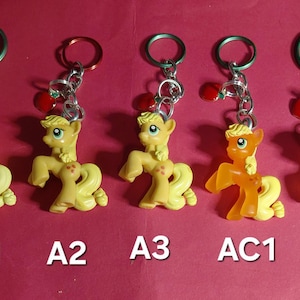 May include: Five keyrings featuring a yellow pony figure with a red apple charm. The pony has a yellow mane and tail, and a red apple mark on its flank. Each keyring has a silver chain and a metal ring.