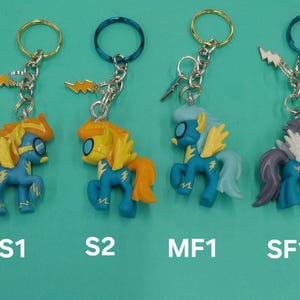 K&ouml;nnte beinhalten: Vier Schl&uuml;sselanh&auml;nger mit Cartoon-Ponyfiguren in Blau, Gelb und Orange. Jeder Schl&uuml;sselanh&auml;nger hat einen Metallring und einen Blitz-Anh&auml;nger. Die Figuren haben unterschiedliche Haar- und Fl&uuml;gelfarben. Der Text "S1", "S2", "MF1" und "SF1" steht darunter.