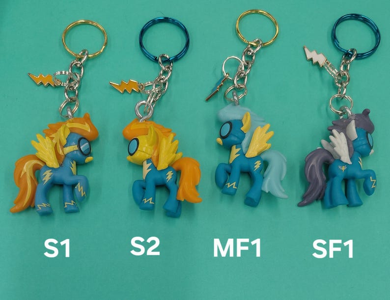 K&ouml;nnte beinhalten: Vier Schl&uuml;sselanh&auml;nger mit Cartoon-Ponyfiguren in Blau, Gelb und Orange. Jeder Schl&uuml;sselanh&auml;nger hat einen Metallring und einen Blitz-Anh&auml;nger. Die Figuren haben unterschiedliche Haar- und Fl&uuml;gelfarben. Der Text "S1", "S2", "MF1" und "SF1" steht darunter.
