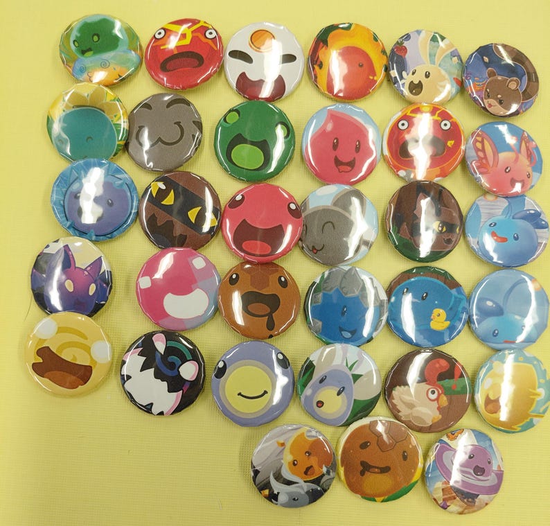 May include: A collection of colorful, round button pins with various cartoon character designs. The pins feature a variety of colors, including green, red, blue, and yellow, and are arranged on a yellow surface. Each pin has a unique, detailed illustration.