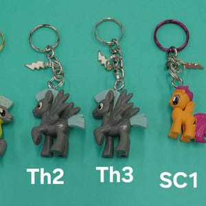 K&ouml;nnte beinhalten: Schl&uuml;sselanh&auml;nger mit bunten Cartoon-Ponyfiguren. Die Schl&uuml;sselanh&auml;nger haben silberne Ketten und Metallringe. Die Ponys sind in verschiedenen Farben, darunter Grau, Orange und Gelb, mit Fl&uuml;geln und anderen Details. Der Text "Th1", "Th2", "Th3", "SC1" und "ChS1" ist unten aufgedruckt.