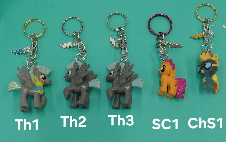 K&ouml;nnte beinhalten: Schl&uuml;sselanh&auml;nger mit bunten Cartoon-Ponyfiguren. Die Schl&uuml;sselanh&auml;nger haben silberne Ketten und Metallringe. Die Ponys sind in verschiedenen Farben, darunter Grau, Orange und Gelb, mit Fl&uuml;geln und anderen Details. Der Text "Th1", "Th2", "Th3", "SC1" und "ChS1" ist unten aufgedruckt.