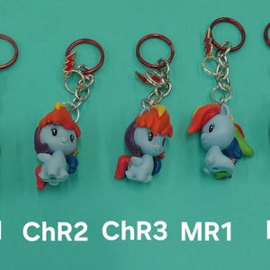 K&ouml;nnte beinhalten: F&uuml;nf Schl&uuml;sselanh&auml;nger mit bunten Cartoon-Ponyfiguren mit roten, orangefarbenen und blauen Haaren. Jeder Schl&uuml;sselanh&auml;nger hat eine silberne Kette und einen roten Ring. Der Text "ChR1", "ChR2", "ChR3", "MR1" und "HR1" ist darunter gedruckt.
