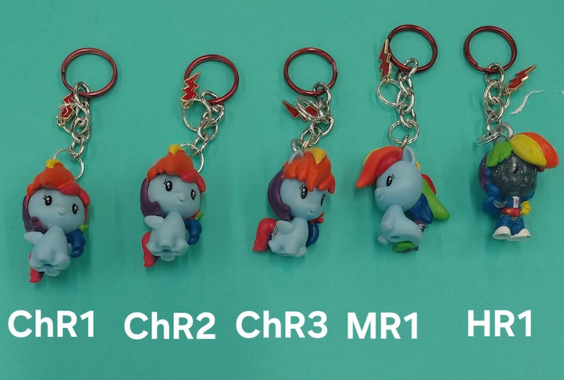 K&ouml;nnte beinhalten: F&uuml;nf Schl&uuml;sselanh&auml;nger mit bunten Cartoon-Ponyfiguren mit roten, orangefarbenen und blauen Haaren. Jeder Schl&uuml;sselanh&auml;nger hat eine silberne Kette und einen roten Ring. Der Text "ChR1", "ChR2", "ChR3", "MR1" und "HR1" ist darunter gedruckt.