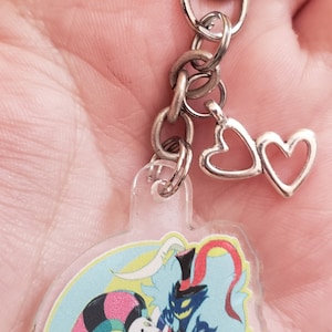 Puede incluir: Un llavero de acrílico transparente con una ilustración de dibujos animados de dos personajes, uno con un traje de bufón y el otro con cuernos y una cola roja, sobre un fondo azul y amarillo. El llavero tiene una cadena plateada y dos dijes de corazón plateados.