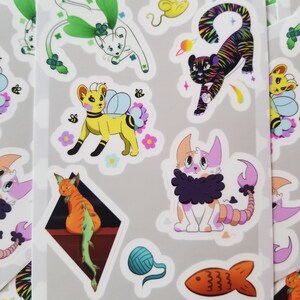 May include: A sheet of colourful cat stickers featuring a variety of designs, including a cat with wings, a cat with a fish tail, a cat with a bee costume, and a cat with a tiger stripe pattern. The stickers are on a grey background with the text "Cat Stickers" at the bottom.