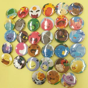 May include: A collection of round, colorful pinback buttons featuring various cartoon character designs. The buttons display a range of colors, including green, red, blue, and brown, with detailed illustrations of creatures and faces. The buttons are arranged closely together.