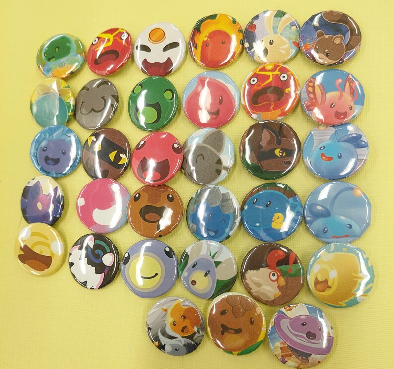 May include: A collection of round, colorful pinback buttons featuring various cartoon character designs. The buttons display a range of colors, including green, red, blue, and brown, with detailed illustrations of creatures and faces. The buttons are arranged closely together.