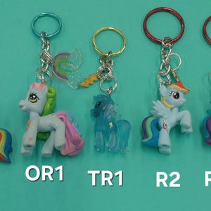 May include: Five colorful My Little Pony keychains. Each keychain features a small pony figure with a rainbow mane and tail, attached to a metal ring. The keychains have different colored rings and charms.