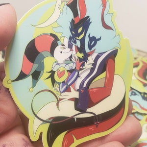 May include: A sticker featuring two cartoon characters, one in a red and black jester costume and the other in a black and purple suit with a red top hat. The jester is holding a heart-shaped object and the other character has a devilish grin. The background is a light blue and yellow gradient.