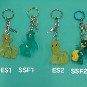 K&ouml;nnte beinhalten: Vier Schl&uuml;sselanh&auml;nger mit Cartoon-Ponyfiguren in Gelb und T&uuml;rkis. Jeder Schl&uuml;sselanh&auml;nger hat einen Metallring und einen Blitz-Anh&auml;nger. Die Schl&uuml;sselanh&auml;nger sind mit ES1, SSF1, ES2 und SSF2 beschriftet.