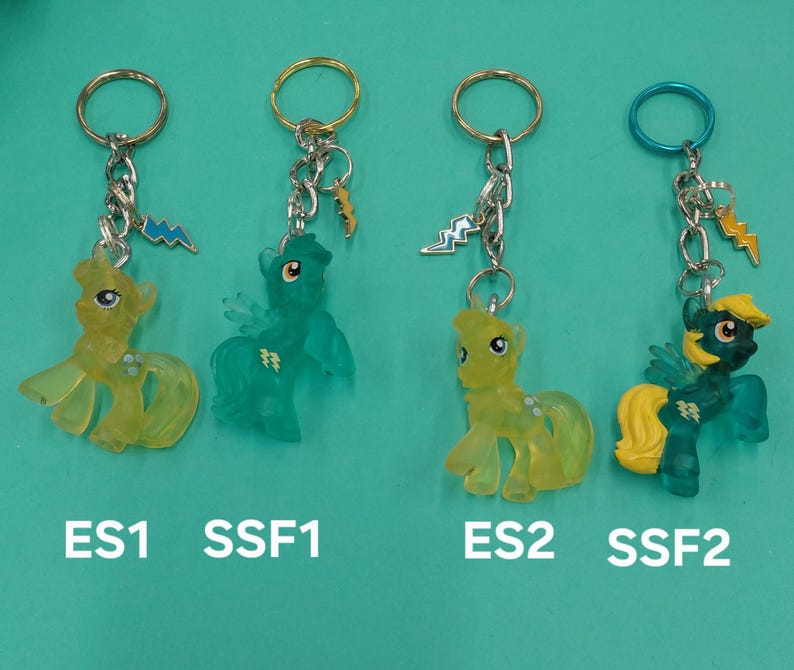 K&ouml;nnte beinhalten: Vier Schl&uuml;sselanh&auml;nger mit Cartoon-Ponyfiguren in Gelb und T&uuml;rkis. Jeder Schl&uuml;sselanh&auml;nger hat einen Metallring und einen Blitz-Anh&auml;nger. Die Schl&uuml;sselanh&auml;nger sind mit ES1, SSF1, ES2 und SSF2 beschriftet.