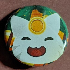 May include: A round button featuring a cartoon cat face with a gold halo. The cat has a white face, black eyes, and a red mouth. The background is a mix of green, orange, and brown.