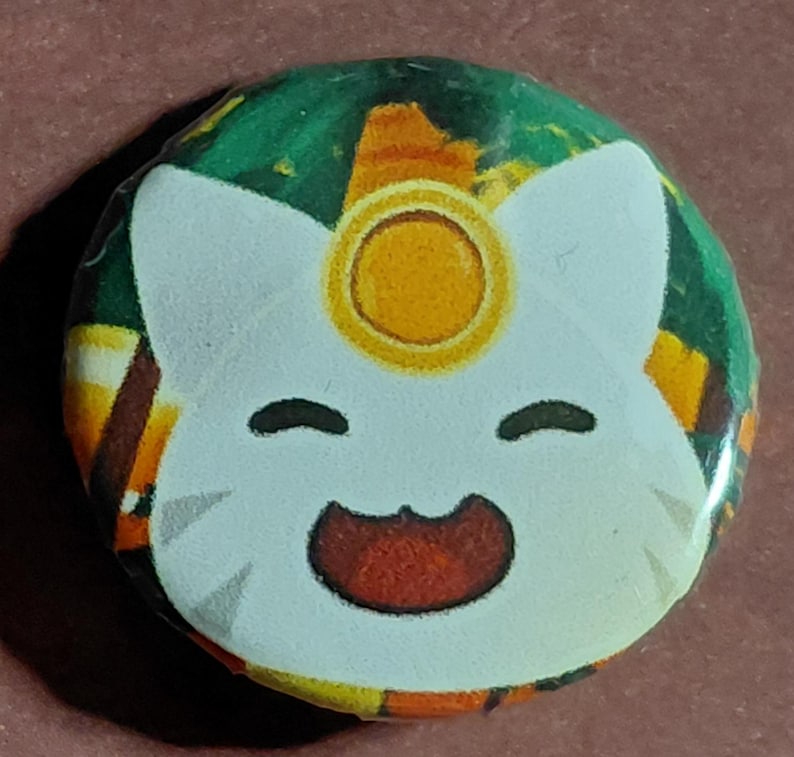 May include: A round button featuring a cartoon cat face with a gold halo. The cat has a white face, black eyes, and a red mouth. The background is a mix of green, orange, and brown.