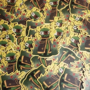May include: A pile of green, brown, and yellow stickers featuring a cartoon character with a green head, brown body, and yellow accents. The character is wearing a brown robe with a gold trim and a yellow sash. The stickers are surrounded by yellow, green, and red flowers.