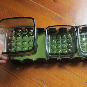 Op de afbeelding: Een vintage serveerblad met vier vierkante glazen schalen. Elke schaal heeft een donkergroene kleur en een patroon van verhoogde stippen. De lade heeft een groene basis en een donkerbruine, geweven rand.