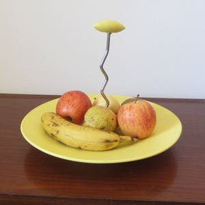 May include: A yellow ceramic fruit bowl with a metal stand and a banana, two red apples, and two yellow pears.