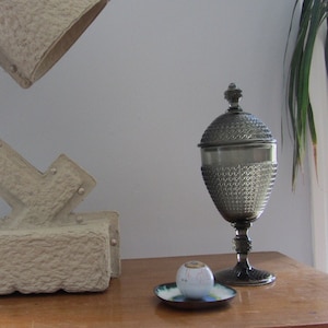 Puede incluir: Una lámpara blanca texturizada con un diseño geométrico se encuentra sobre una mesa de madera junto a un frasco de vidrio decorativo plateado con tapa.