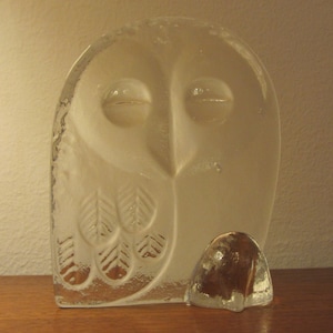 Pode incluir: Uma escultura de coruja de vidro transparente com um design detalhado de penas e olhos. A coruja está em pé sobre uma base de vidro redonda menor.