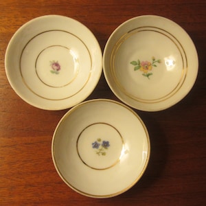 Może przedstawiać: Trzy małe białe porcelanowe talerzyki z pozłacanymi brzegami i kwiatowymi wzorami. Talerzyki mają pojedynczy kwiat w środku, każdy w innym schemacie kolorystycznym.