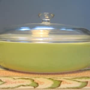 Può includere: Una pirofila in vetro verde vintage con un coperchio in vetro trasparente. La pirofila ha una forma arrotondata e una superficie liscia.