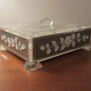 Puede incluir: Una caja de plástico transparente con una base gris y un diseño floral. La caja tiene una tapa con bisagras y se apoya en cuatro patas de plástico transparente.